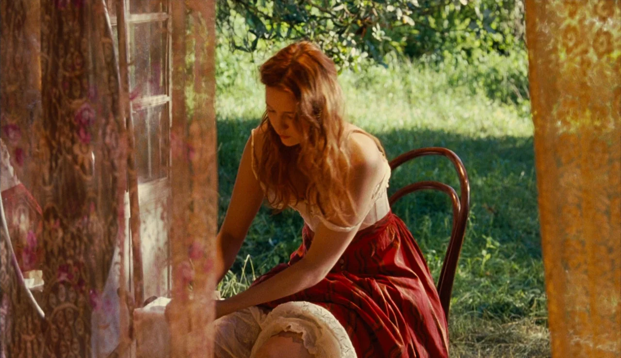 Renoir (2012) movie still: medium — A woman sits in a chair on a veranda edge facing a grassy area. She is working on a piece of fabric while…