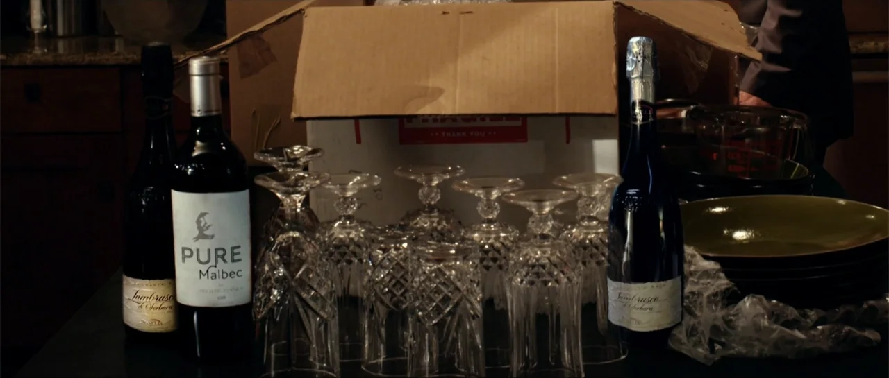 Rage (2014) movie still: medium — A dark countertop holds multiple crystal glasses and wine bottles in front of a box. An opened cardboard box sits…