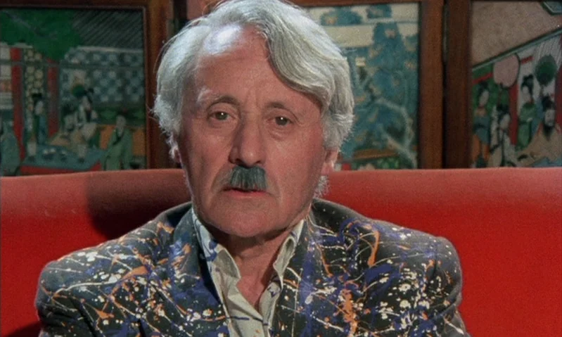Jubilee (1978) movie still: closeup — An older man sits before a folding screen with traditional artwork. He faces the camera directly while wearing…