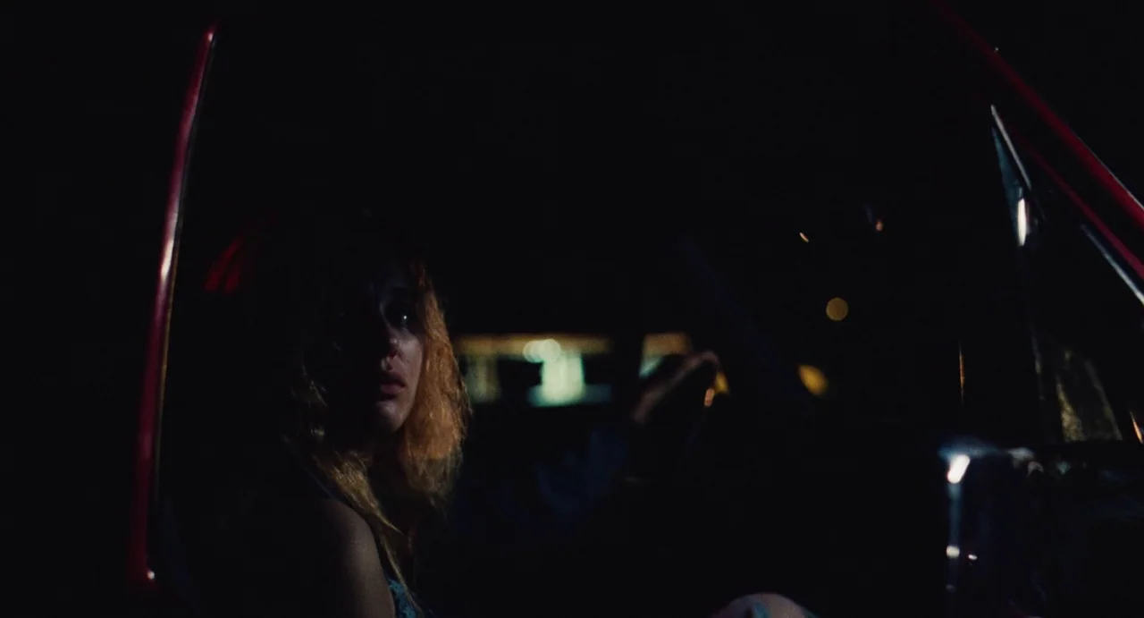 The Giant (2019) movie still: medium — A dark, cramped interior of a vehicle at night with red painted metal door frame. A young woman with wavy…