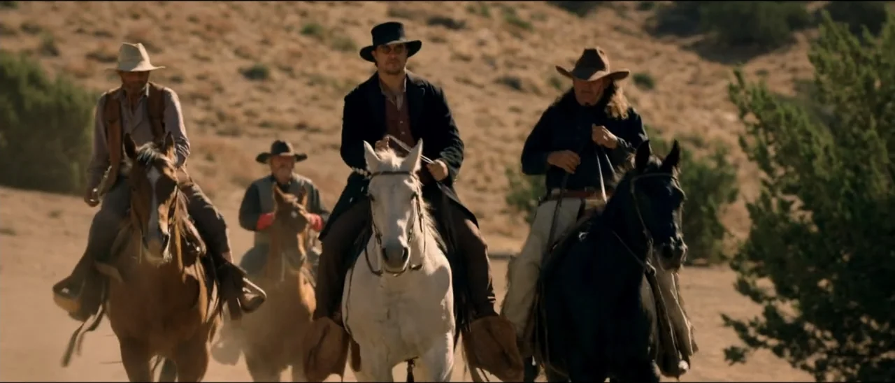 Dead for a Dollar (2022) movie still: fullbody — Four men ride horses across a dry dusty terrain under bright daylight. The group approaches the…