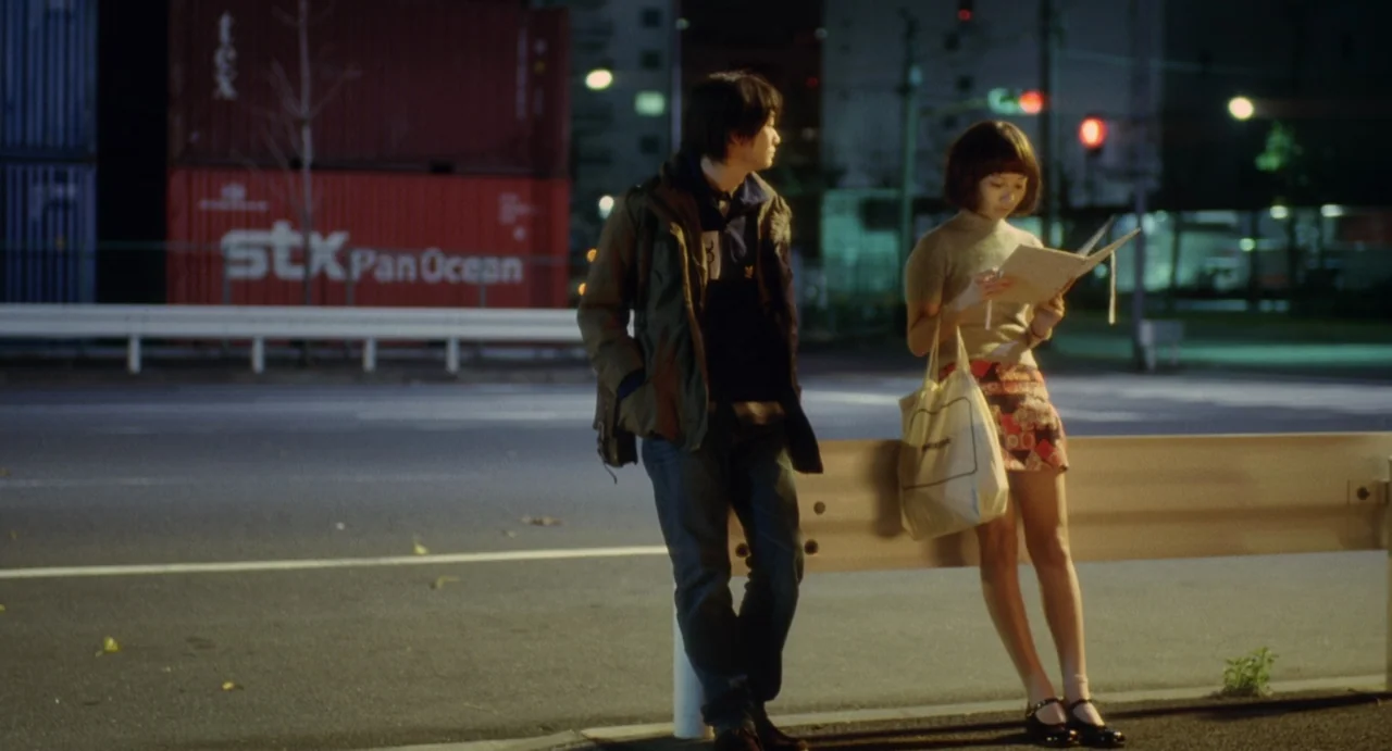 Air Doll (2009) movie still: fullbody — An asphalt road at night with large red shipping containers in the background. A man and a woman stand by a…
