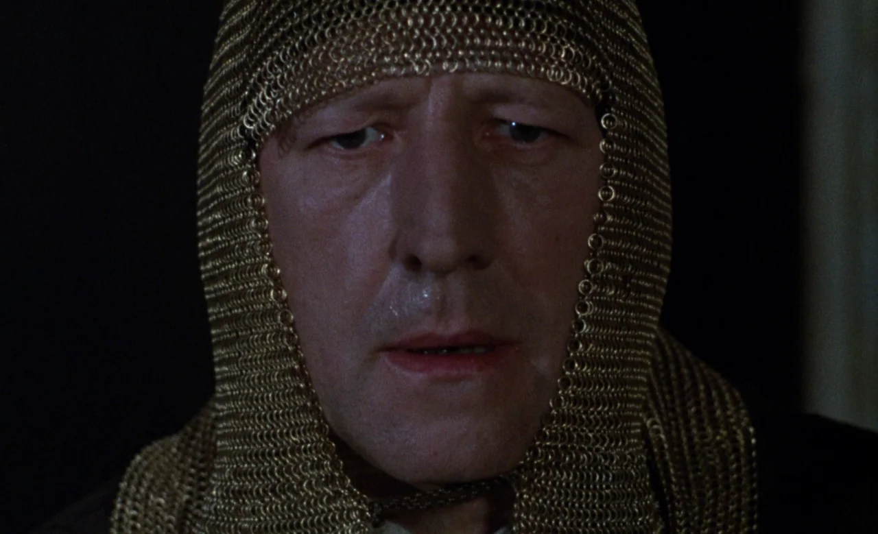 Blanche (1971) movie still: closeup — A dark, nondescript background frames a man wearing a metallic gold chainmail headpiece. The man looks forward…