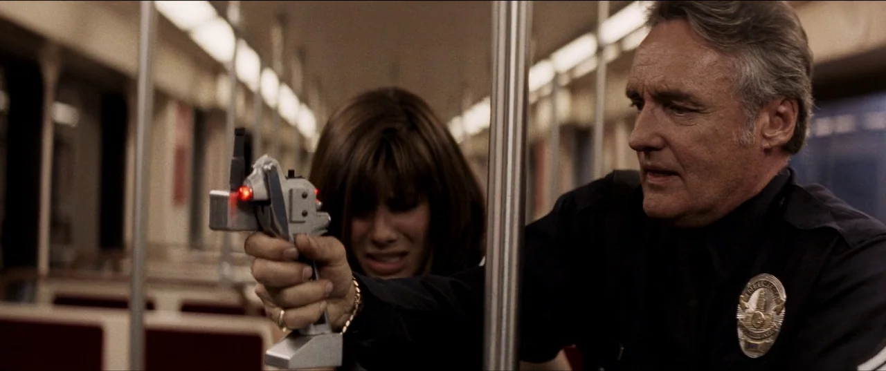 Speed (1994) movie still: medium — The interior of a subway car features rows of seats and metallic support poles. A male police officer aims a…