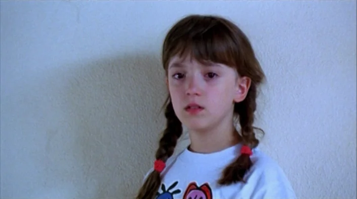Code Unknown (2000) movie still: closeup — A young girl stands against a plain, light-colored wall in a simple room. She stares slightly off-camera…