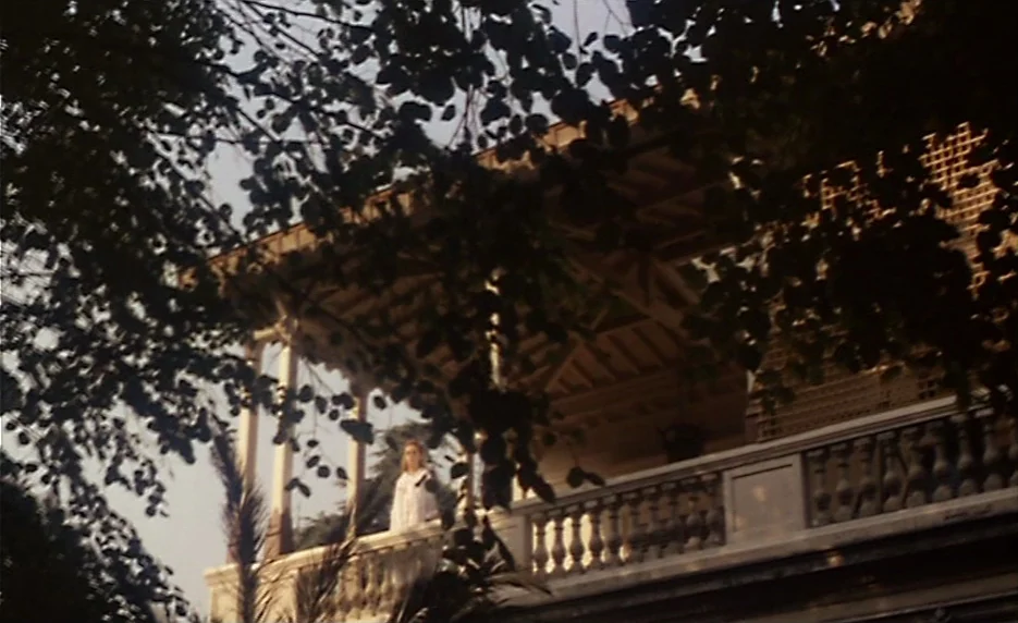 The Immortal Story (1968) movie still: wide — A classic veranda with stone balusters sits partially obscured by lush, overhanging tree branches. A…