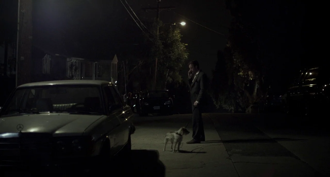 Beginners (2010) movie still: wide — A quiet residential road at night is illuminated by a single overhead streetlamp. A man in a suit stands on the…