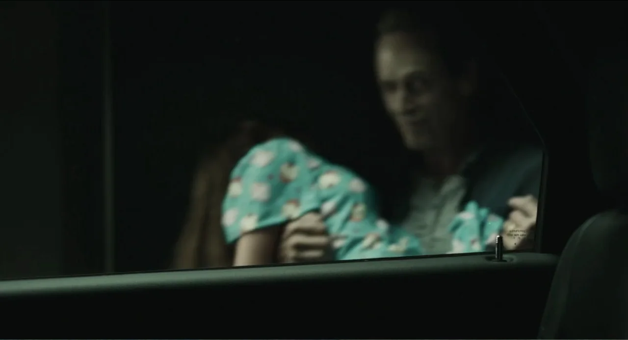 Haunter (2013) movie still: medium — A dark night exterior viewed through the side window frame of a car. An adult man holds a child close while…