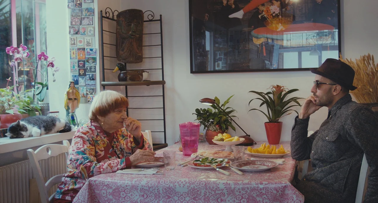 Faces Places (2017) movie still: wide — A sunlit room with a dining table featuring a pink patterned tablecloth and food. An elderly woman and a man…