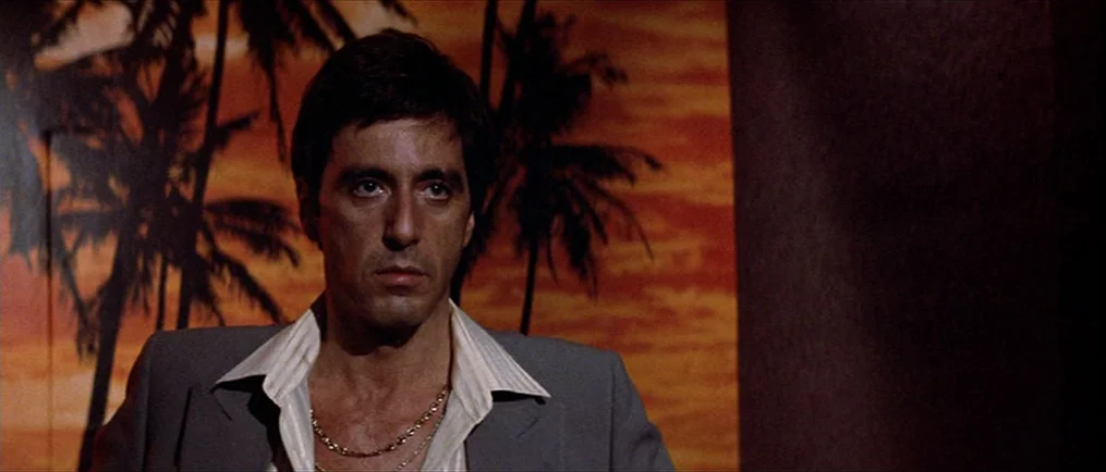 Scarface (1983) movie still: medium — A dimly lit room features a mural of a tropical sunset with palm trees. A man stands in the foreground wearing…