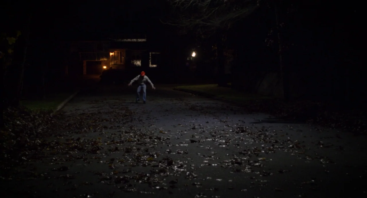 Happer’s Comet (2022) movie still: wide — A dark residential street is covered in dry brown autumn leaves at night. A lone person wearing a red hat…