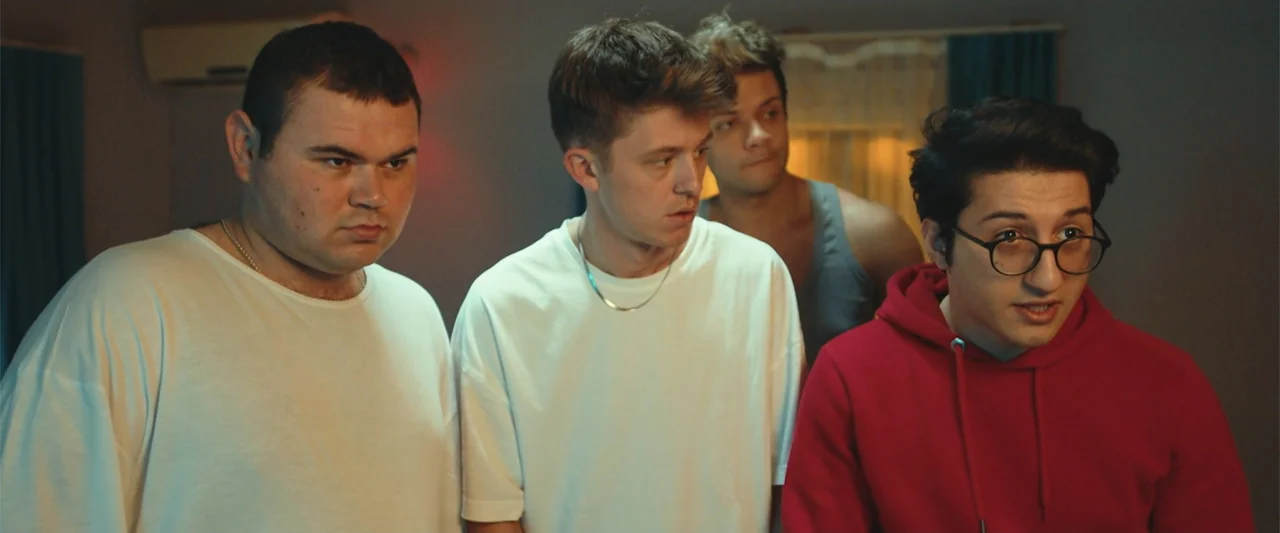 Wrong Package (2025) movie still: medium — A dimly lit room provides the backdrop for three men standing closely together. The subjects look…