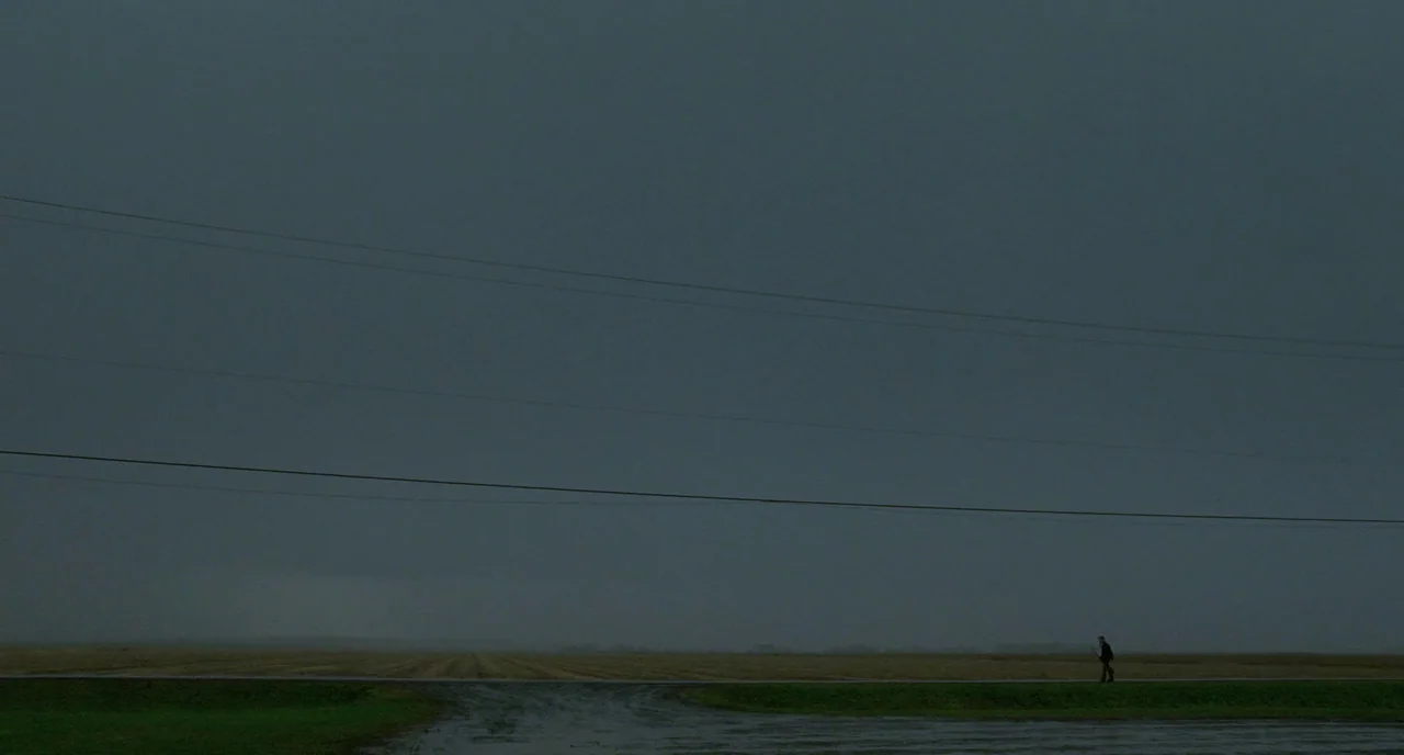Tom at the Farm (2013) movie still: establishing — A vast, flat, overcast rural landscape with a thin layer of clouds above. A lone figure walks…