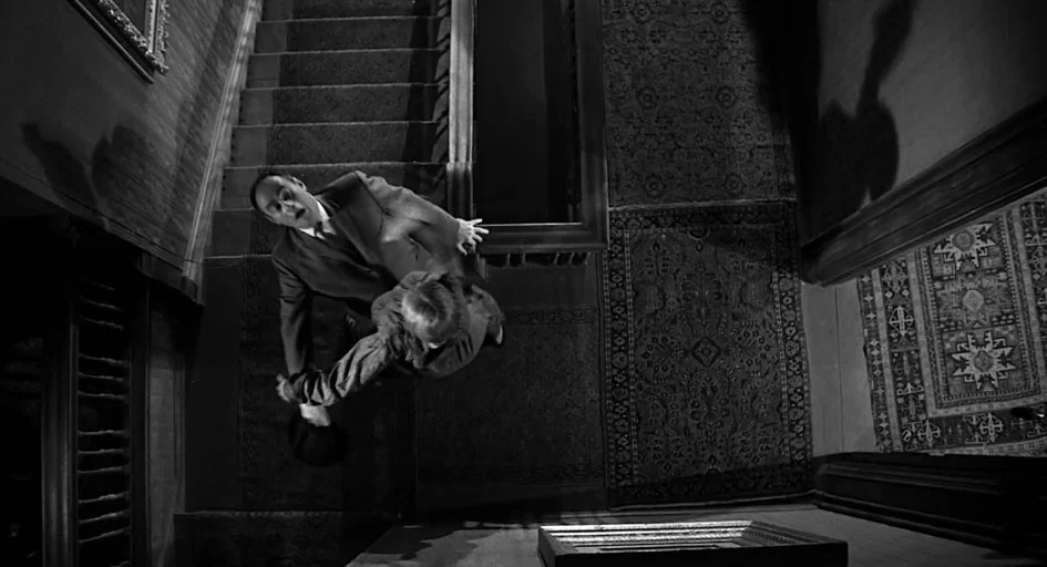 Psycho movie still: wide — A dark, carpeted wooden staircase extends upward from a landing in a period house. A man and a woman hold onto each other…