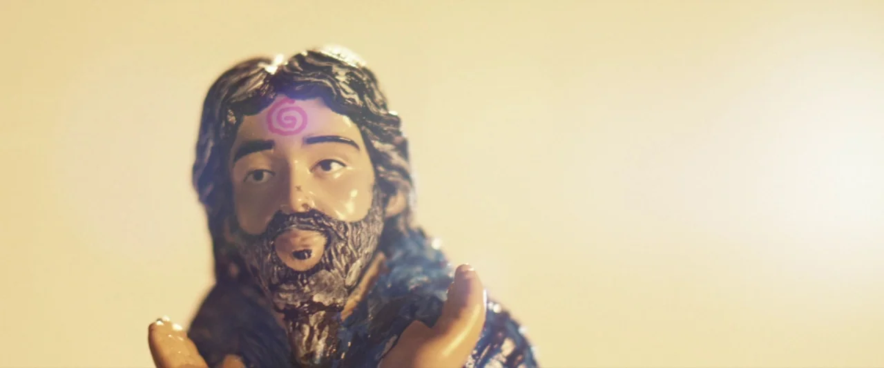 Seven Stages to Achieve Eternal Bliss (2018) movie still: closeup — A close-up view of a painted ceramic male figurine against a plain beige…