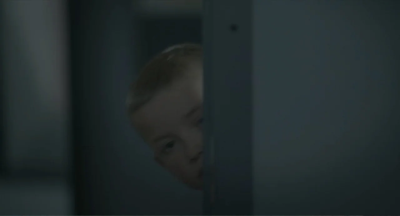 Orphan: First Kill (2022) movie still: closeup — A dark interior space features a vertical wooden frame blocking part of the view. A young boy looks…