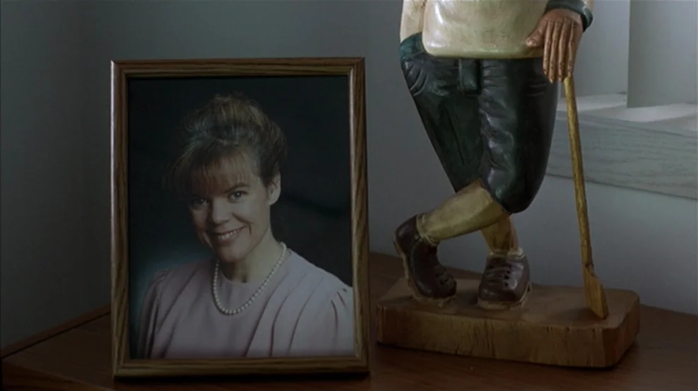 Fargo (1996) movie still: medium — A wooden tabletop displays a framed portrait of a woman next to a figurine. The carved wooden figurine depicts a…