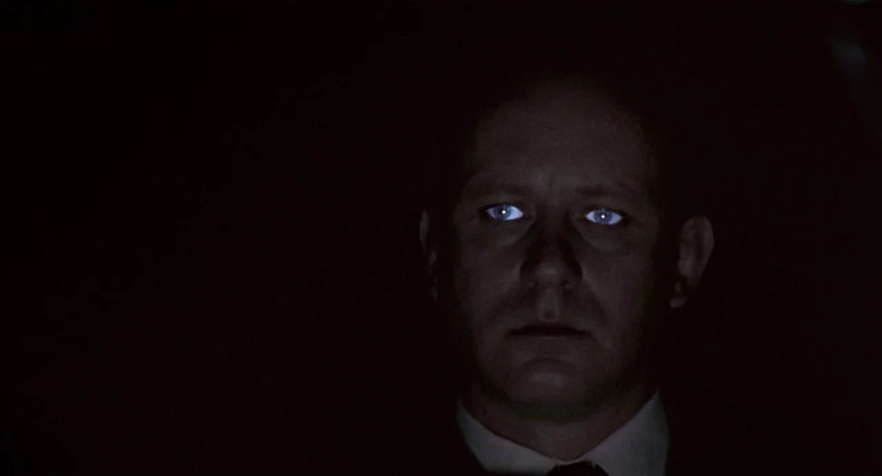 Insomnia (1997) movie still: closeup — A dark vehicle cabin at night with total shadow surrounding the subject. The man looks forward as blue light…