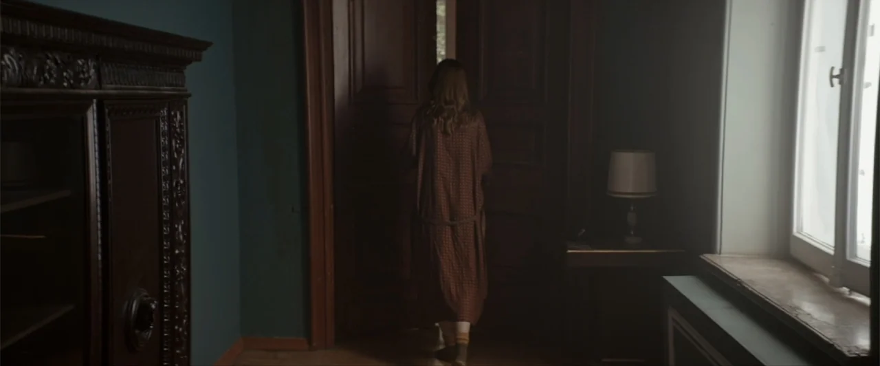 La herencia (2010) movie still: fullbody — A dark, moody room with teal walls, a tall wooden wardrobe, and a side window. A woman wearing a dressing…