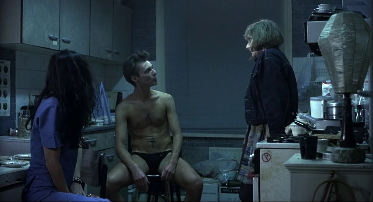 Naked (1993) movie still: medium — A dimly lit room with cluttered kitchen cabinets and a central stove. A man sits shirtless on a small stool while…
