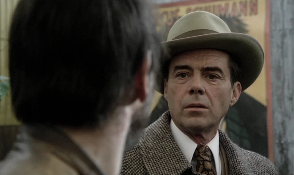 Despair (1978) movie still: medium — A man in a tweed blazer and fedora stands outdoors near a colorful poster. The man looks at an unseen person…
