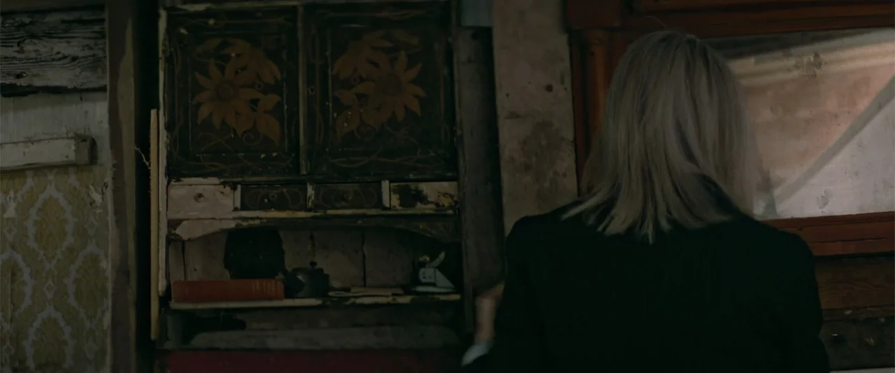 Hampstead (2017) movie still: medium — An interior space features aged walls with peeling vintage wallpaper and a dark wooden cabinet. A woman with…