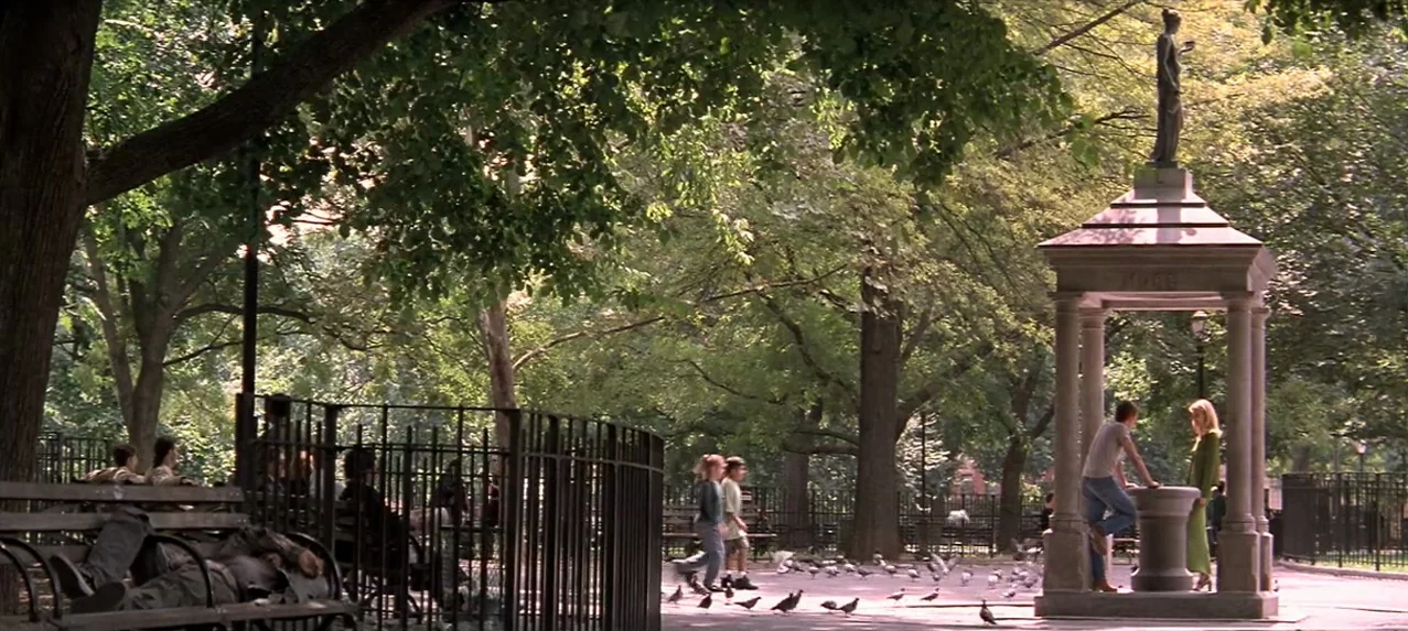 Great Expectations (1998) movie still: wide — A sunny public park features mature trees and a stone fountain structure with a statue. A couple…