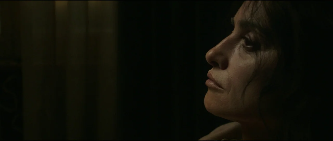 Ferrari (2023) movie still: closeup — A dimly lit, shadowed space provides a stark backdrop for the woman. A middle-aged woman appears in profile…