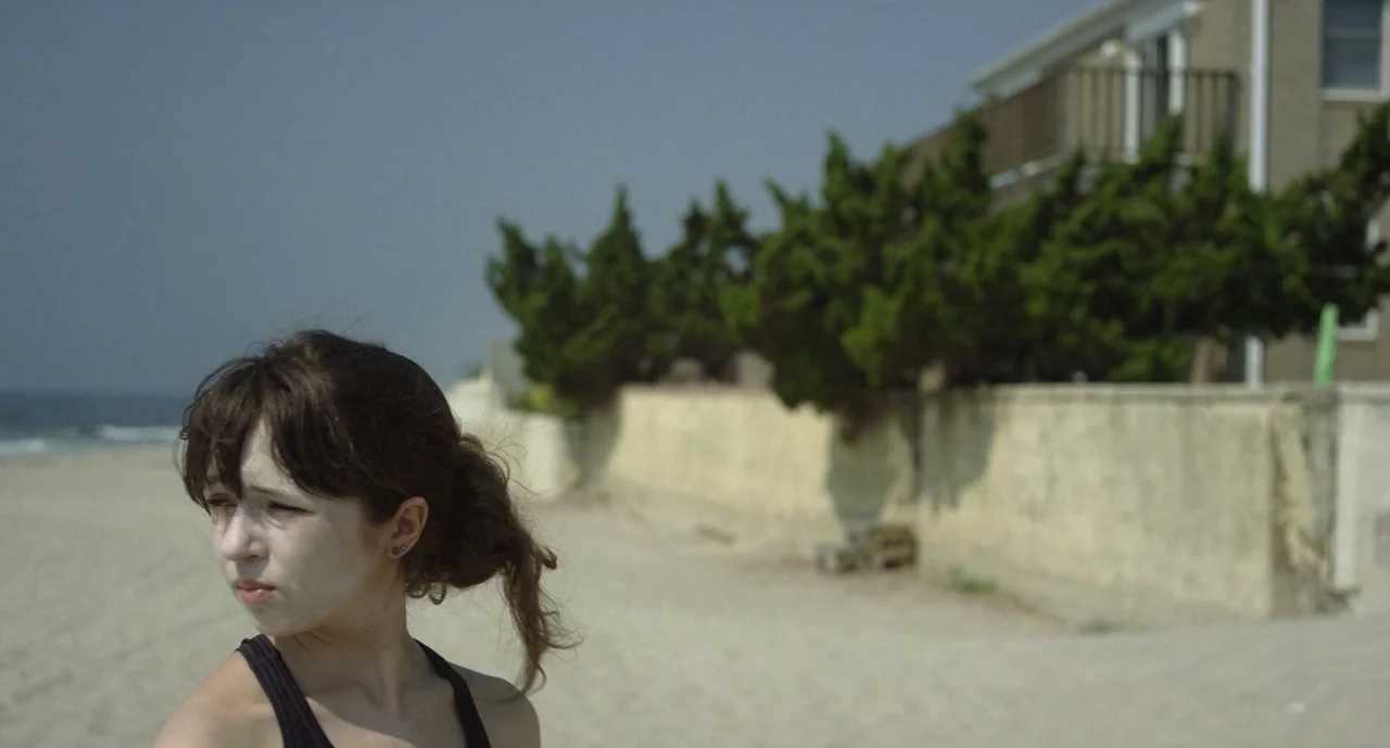 It Felt Like Love (2013) movie still: medium — A sandy beach stretches toward a retaining wall topped with green evergreen bushes. A woman with…