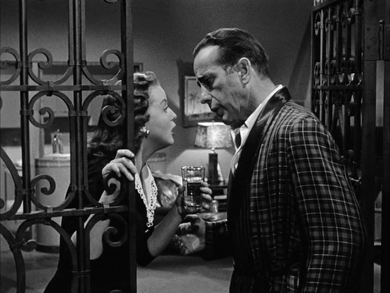 In a Lonely Place (1950) movie still: medium — A man and a woman converse while separated by an intricate metal security gate. The man wears a plaid…