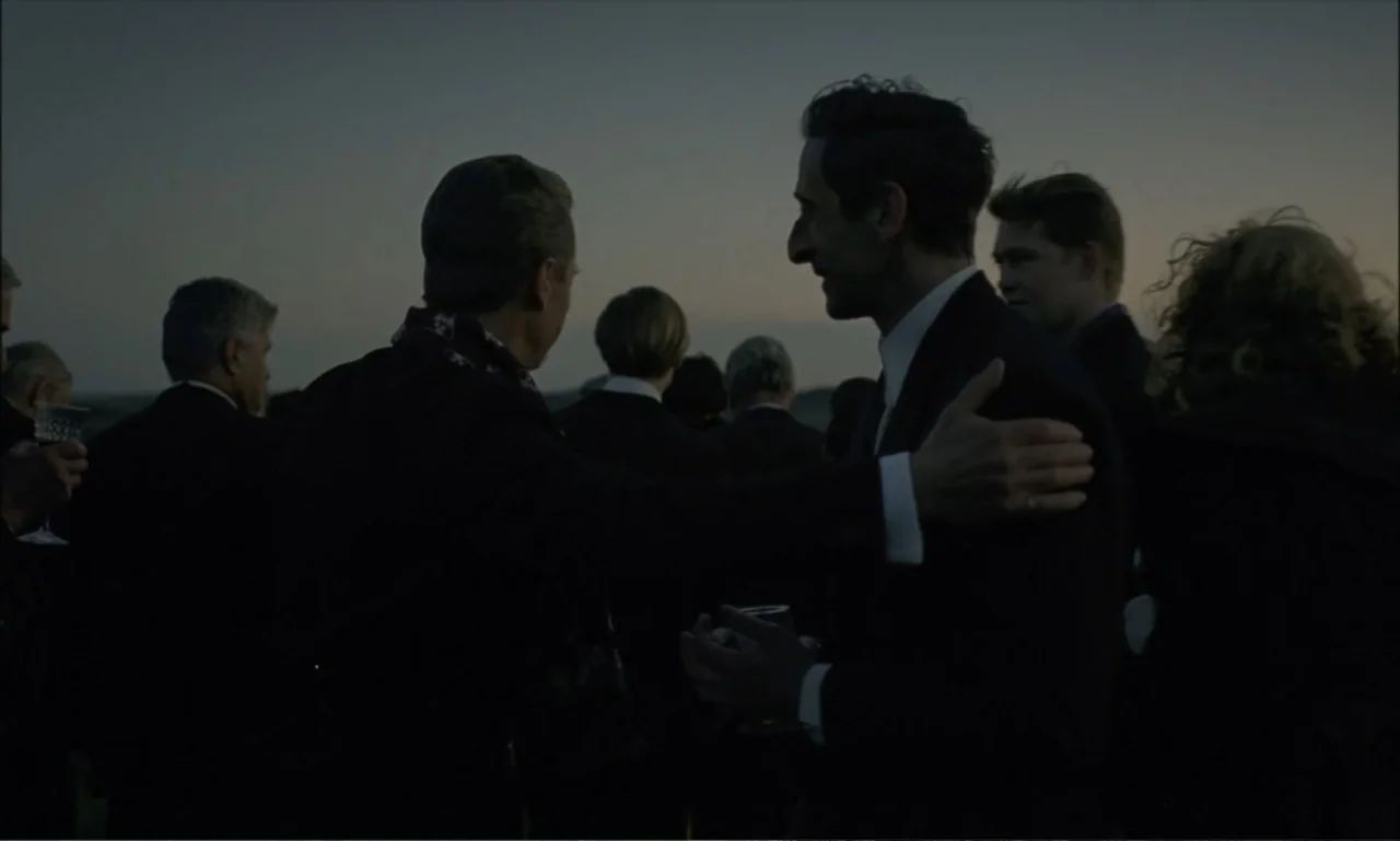 The Brutalist (2024) movie still: medium — A group of people gathers outdoors at dusk under a dim evening sky. One man turns to greet another man by…