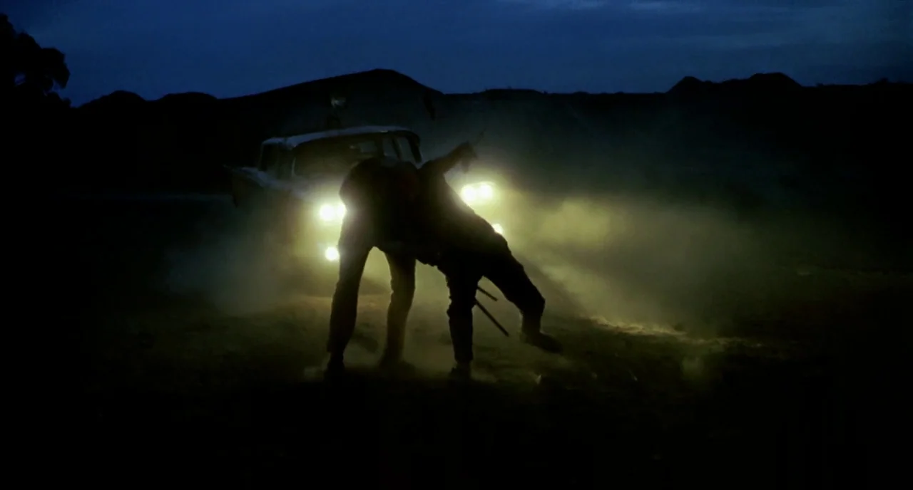 Wake In Fright (1971) movie still: wide — A dark, desolate desert landscape features a parked vintage car at night. Two men are locked in a physical…