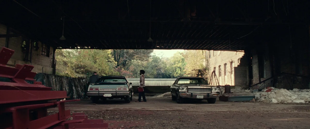 BlacKkKlansman (2018) movie still: wide — A dark, dilapidated open-air industrial structure reveals an exterior view of trees and water. Two men…