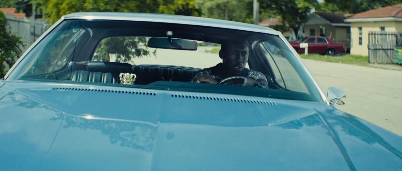 Moonlight (2016) movie still: medium — A light blue car moves down a quiet residential street with single-story homes. The driver is focused ahead…