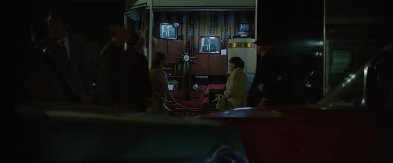 Reagan (2024) movie still: wide — A night view of a 1950s storefront display window filled with several CRT televisions. Several people stand on the…