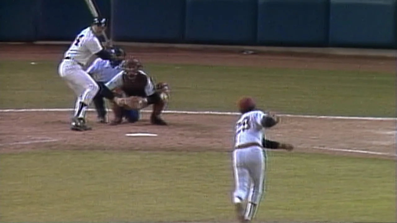 Reggie (2023) movie still: wide — A baseball diamond during a game with a pitcher throwing toward home plate. The pitcher is mid-motion while the…