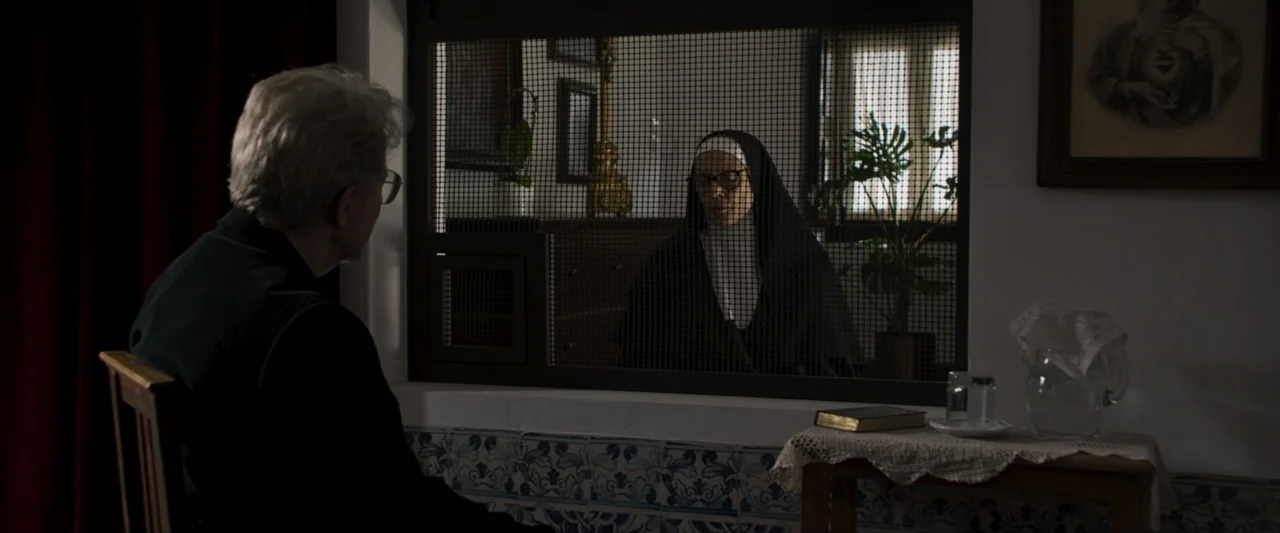 Fatima (2020) movie still: medium — An interior space features a partition window with a wire grid between two individuals. One subject sits in…