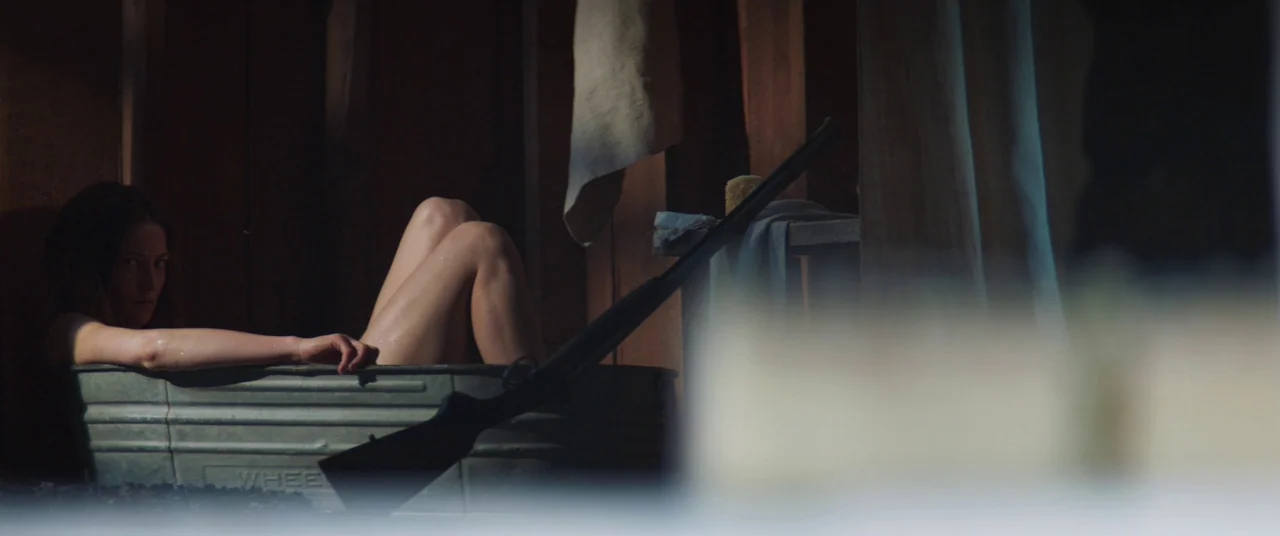 The Wind (2018) movie still: medium — A rustic wooden washroom with rough-hewn boards and a metal tub. A woman sits in the tub while a rifle rests…