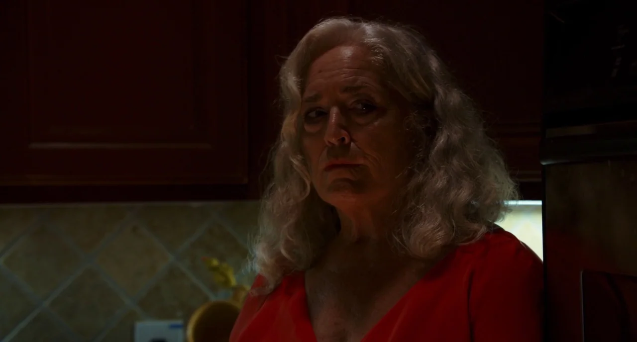 Krisha (2015) movie still: medium — A dimly lit home environment featuring dark wooden cabinetry and light tiled backsplash. An older woman with…
