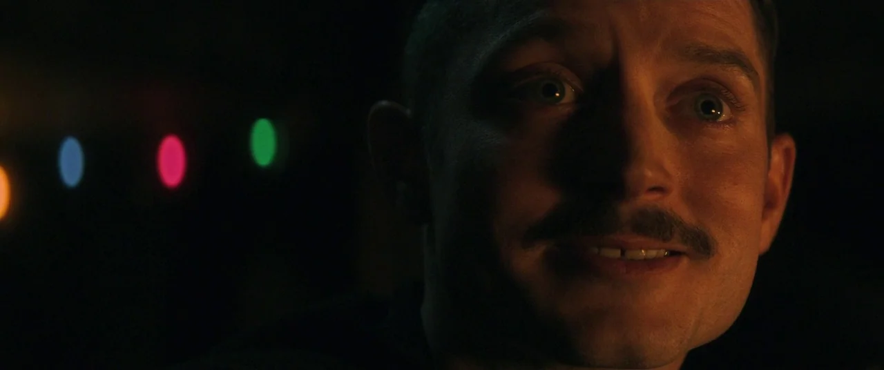 Come To Daddy (2019) movie still: closeup — A dark bar interior features blurred multicolored string lights against a black backdrop. A man with a…