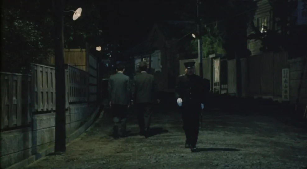 Madadayo (1993) movie still: fullbody — A narrow, unpaved urban road is lined with high wooden fences at night. Two men walk away from the viewer…