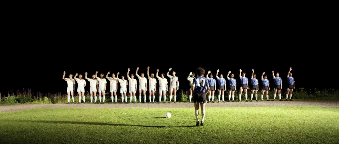 Youth (2015) movie still: wide — A soccer field at night features a line of players standing against a black background. One player in a blue jersey…