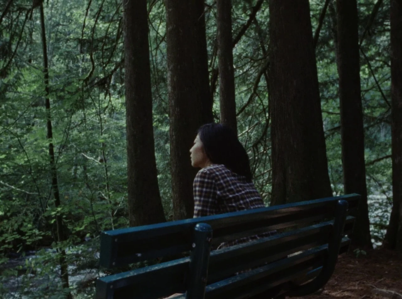 Riceboy Sleeps (2022) movie still: medium — A dense forest environment with thick tall trees behind a dark wooden bench. A young woman sits on the…