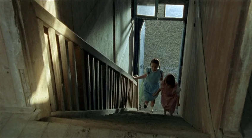 Bluebeard (2009) movie still: wide — A dark, wood-paneled staircase leads down to an open doorway showing a pebbled outdoor surface. Two young girls…