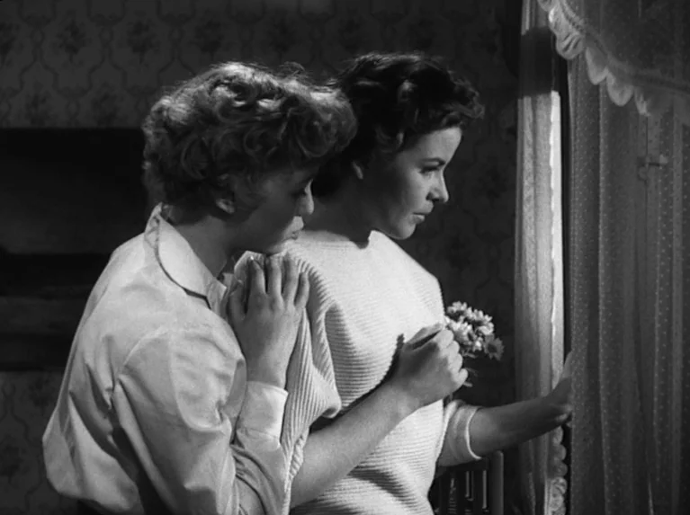 Waiting Women (1952) movie still: medium — Two women stand together in a dimly lit room with patterned wallpaper. One woman looks out a window while…