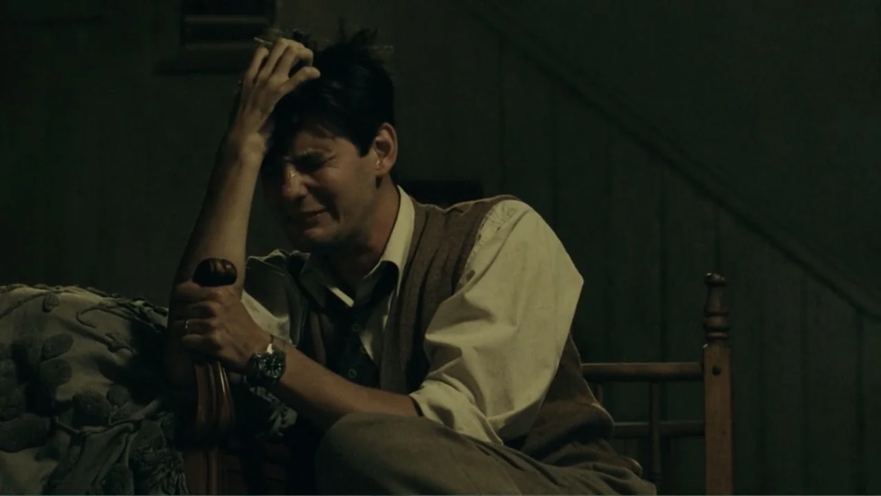 The Words (2012) movie still: medium — A dark, spartan bedroom contains a wooden headboard and dim lighting. A young man sits on the bed edge while…