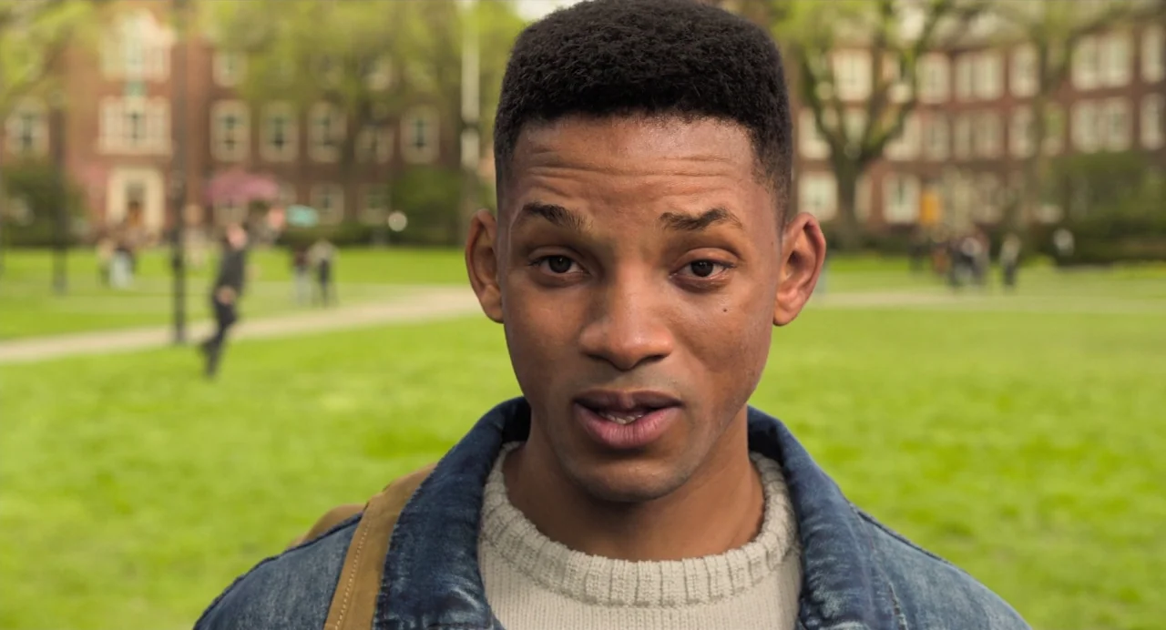 Gemini Man (2019) movie still: closeup — A grassy university campus quad sits before a classic red brick building. A young man stands in the…