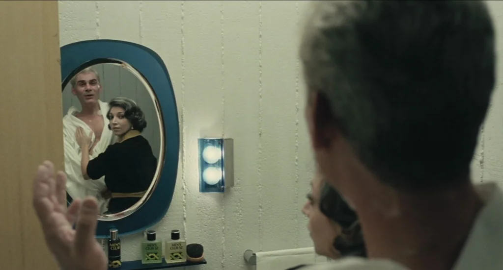 Todo Modo (1976) movie still: medium — A modern 1970s bathroom features textured off-white walls and a blue-framed oval mirror. A woman stands…