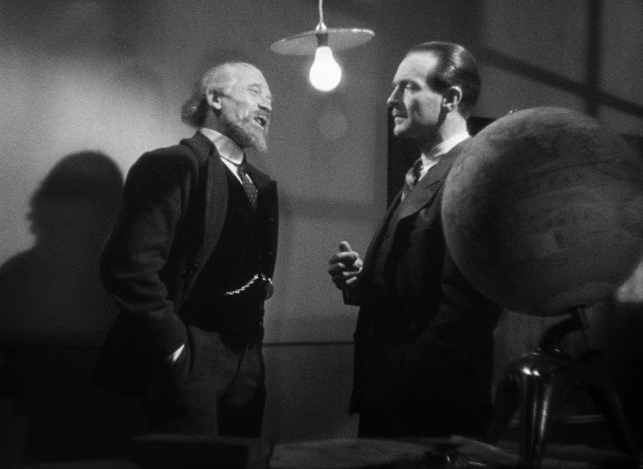 Le Corbeau (1943) movie still: medium — Two men stand in a dimly lit, shadow-filled room under an exposed lightbulb. The older bearded man faces a…