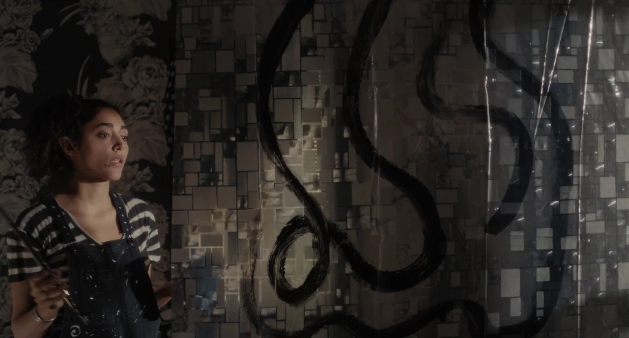 Paterson (2016) movie still: medium — A dark space features patterned wallpaper on one side and a plastic sheet surface. The woman stands holding a…